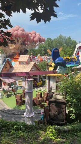 The video shows a whimsical amusement park scene with brightly colored houses, cartoonish characters, and whimsical decorations. In the foreground, a large purple and white striped pole stands. Behind it, a small train track curves through a village with small houses, complete with potted plants. A yellow tractor is parked near a building with a blue door. In the background, a large, tree-like structure is adorned with numerous pink balloons. A windmill with a yellow roof is also visible. The scene is set on a sunny day with green grass and lush trees.