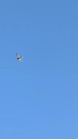 A small, single-engine airplane is captured in flight against a clear blue sky. The aircraft, a low-wing monoplane, appears to be white with black and orange accents or markings. Its propeller is visible, and it is angled slightly upwards, suggesting it is climbing or maneuvering. The plane is positioned in the upper left quadrant of the frame. The vast expanse of the cloudless sky dominates the scene, indicating a bright, sunny day. The image was taken from a ground-level perspective, looking up at the aircraft. There is no visible text within the frame. The setting is likely an outdoor location with open skies, fitting for aviation activities. The overall mood is serene and captures a moment of flight.