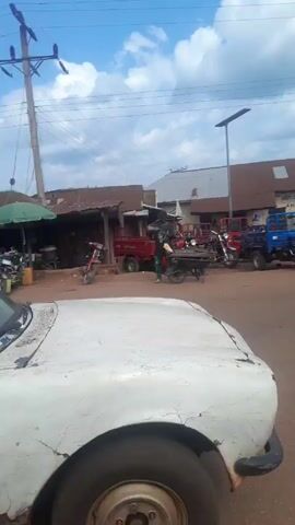 The video shows a busy street scene in a town or village in what appears to be an African country. The sky is blue with scattered clouds.  In the foreground, the side of a white car is visible.  Further down the street, several three-wheeled vehicles and motorcycles are parked. People are walking about and milling around.  A white pickup truck drives by, its bed filled with women wearing matching maroon and green patterned outfits. A young man stands in the back of the truck as it moves. A man on a motorcycle rides past the camera. The buildings along the street appear to be simple, with metal roofs.
