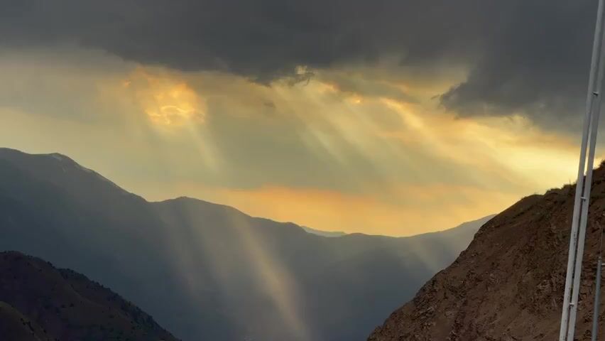 This video captures a majestic mountain landscape bathed in dramatic sunlight streaming through dark, cloudy skies. The sun's rays, known as crepuscular rays, pierce through the atmosphere, creating ethereal beams of light that illuminate the valleys and slopes. The mountains themselves are layered, with distant peaks fading into a soft haze, suggesting a vast and expansive terrain. The foreground features a rugged, rocky incline on the right side, contrasting with the softer, more vegetated slopes on the left. The overall atmosphere is one of awe-inspiring natural beauty, with the interplay of light and shadow emphasizing the raw power and grandeur of the mountainous environment. There are no people, animals, or explicit man-made structures visible, focusing solely on the natural spectacle.