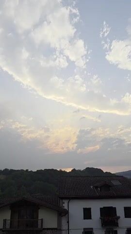 This vertical video captures a serene sunset in Baztan, Spain. The sky is a prominent feature, filled with a dramatic display of clouds. A large, wispy cloud formation stretches horizontally across the upper portion of the frame, illuminated by the setting sun. Below this, a more textured layer of clouds reflects softer hues of yellow and orange, hinting at the fading daylight.

The scene transitions to a rustic village setting. In the foreground, the tiled roofs of traditional houses are visible. The houses appear to be made of stone and white plaster, with dark shutters on the windows. One house features a small balcony with a railing. A hint of greenery, possibly from flowering plants, adorns the balcony. Behind the houses, a densely forested hillside forms the backdrop, its dark green foliage silhouetted against the lighter sky. The distant hills create a layered effect, fading into the horizon.

The time of day is clearly sunset, indicated by the warm, diffused light and the color palette of the sky. The weather appears calm, with the clouds suggesting a peaceful end to the day rather than any impending storm. The location cues, such as the architecture of the houses and the lush, hilly landscape, strongly point to a rural setting in Baztan, Spain. No people are visible in the frame, contributing to the tranquil and uninhabited feel of the scene. The overall emotion conveyed is one of peace and natural beauty. There is no visible text in the video.