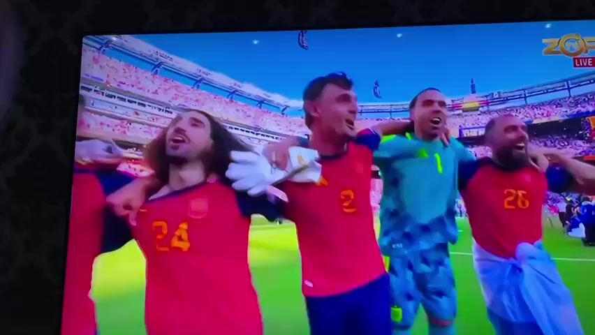 The video displays a television screen showing a soccer match. In the first frame, four male soccer players in red and yellow uniforms are celebrating on a green field, with a stadium and cheering crowd in the background. The second, third, and fourth frames focus on a single male soccer player wearing a light blue and white striped jersey. He is sitting on the grass, head down, with his hand on his knee, appearing to be crying. His face is partially visible and he has tattoos on his arm and neck. The background in these frames is a green grass field. The broadcast appears to be live, indicated by the "LIVE" text in the upper right corner of the screen.
