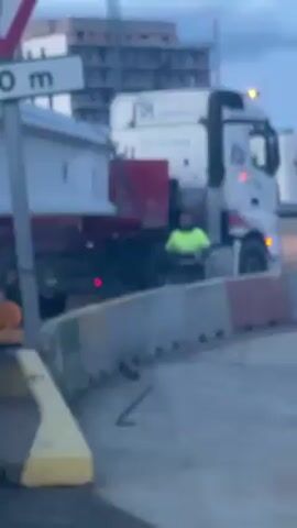 The video shows a man wearing a neon yellow shirt standing next to a large white truck at a construction site. The truck has a red panel on its side and is partially obscured by concrete barriers. There is a road sign indicating a roundabout 100 meters ahead. A building under construction is visible in the background. The camera is somewhat blurry, and the lighting suggests it is either dawn or dusk. The truck is stationary, and the man appears to be observing it. The overall impression is that of a construction or road work project in progress.