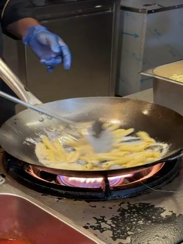 In a commercial kitchen setting, a chef wearing blue gloves and a black uniform mixes penne pasta, a white sauce, and what appears to be blueberries in a large, round wok. The wok sits atop a gas burner, with a visible blue flame. The chef uses a large, silver spoon to stir the mixture. In the background, stainless steel surfaces and equipment are visible, including a storage unit, a container holding pasta, and a red storage unit. The lighting is bright and artificial, highlighting the details of the cooking process.
