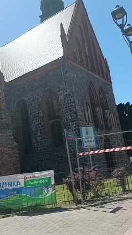 This video captures an outdoor scene featuring a large, old brick building, likely a church, under a clear blue sky. The building's architecture suggests a Gothic style, with pointed arch windows and a steeply pitched roof. 

In the foreground, a banner with text and graphics is visible. The text appears to be in Polish and mentions "PIELGRZYMKA" (pilgrimage) and "Jasna Góra" (a significant pilgrimage site in Poland). Other text on the banner includes dates like "13.08", "26.07", "29.07", and "30.07," indicating that the pilgrimage is a scheduled event. There are also images of people walking, suggesting a journey or movement.

To the right of the banner, a fence encloses an area, and a sign with Polish text, possibly related to the pilgrimage or a local event, is attached to it. A pedestrian crossing sign is prominently displayed on a pole in front of the building, indicating a public area.

The lighting suggests it is daytime, likely late morning or afternoon, with bright sunlight casting shadows. The overall atmosphere seems peaceful and possibly festive, given the context of a pilgrimage. The camera is angled upwards, emphasizing the grandeur of the building against the sky. The video is likely taken in Gryfino, Poland, as indicated by the location cue.