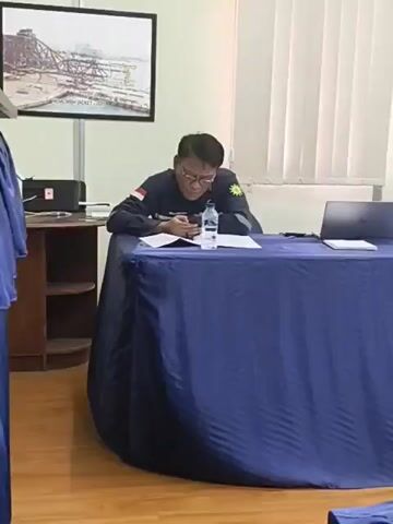 The video shows a meeting room with a long table covered by a blue tablecloth. Several people are seated around the table, most of them with laptops and documents in front of them. The room appears to be an office or conference room, with framed pictures on the walls and blinds on the windows. In the first few seconds, a person in a dark shirt with a red and white armband is seen sitting at the table, looking down at papers. Later, another person in a similar shirt is seen briefly interacting with documents. The subsequent frames show various angles of the meeting in progress, with people engaged in their tasks, some looking at their laptops, others at papers. Bottles of water and cups are also visible on the table. The overall atmosphere seems to be one of focused work or discussion.