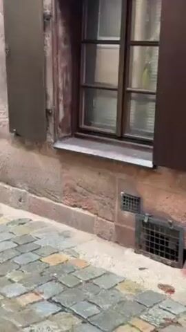 The video shows the exterior of a building. The bottom portion of the building is constructed of reddish-brown stone, while the upper portion has a timber frame with white infill panels. The building has a window with dark wooden shutters, one of which is partially open. A cobblestone street is visible in the foreground.