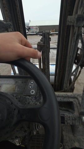 The video shows a close-up, point-of-view shot from inside the cab of a dirty forklift. The viewer is looking out through the forklift's mast, which is composed of multiple vertical metal beams and chains. A hand is visible briefly on the black steering wheel. The instrument panel is dirty and has a few buttons. The ground outside is wet, and a building and some construction materials are visible in the distance. The overall scene appears to be a construction or industrial site.