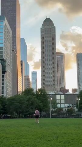 A person wearing a dark t-shirt and shorts is kicking a soccer ball in a grassy park. Several tall buildings, including skyscrapers, are visible in the background, some with sunlight reflecting off their glass facades. The sky is cloudy with patches of sunlight. Trees are also present in the park area. The video appears to be filmed from a distance, capturing a wide view of the urban landscape and the activity in the park.