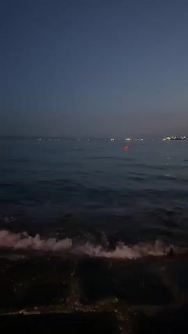 A body of water, likely the sea, is visible in the foreground with gentle waves lapping against a dark, textured surface, possibly a dock or shore. The sky is a deep, twilight blue. In the distance, across the water, numerous small lights can be seen, suggesting a shoreline or boats. One prominent red light stands out on the water. The overall atmosphere is calm and serene, with the fading light of dusk.