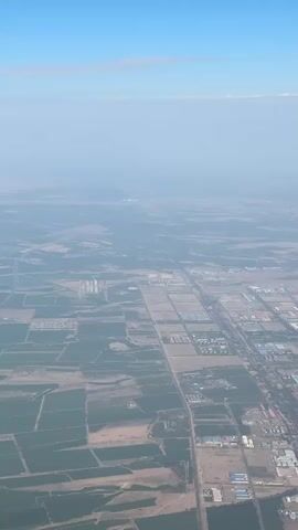 This media file shows an aerial view of a sprawling landscape, likely a rural or semi-urban area in Erliugong, China, due to the presence of agricultural fields interspersed with industrial or commercial buildings. The scene is viewed from a high altitude, suggesting it was captured from an airplane.

The primary subjects are the landscape itself, characterized by a mix of cultivated fields, some appearing harvested and others still green, and clusters of buildings. These buildings vary in size and color, with many appearing to be warehouses or factories, arranged in organized blocks. Roads and pathways crisscross the area, connecting the different sections. In the distance, a hazy horizon suggests a larger settlement or city.

The activity taking place is not dynamic; it's a static, high-altitude observation of the land and its development. There are no people or specific objects of focus visible from this height.

Notable details include the time of day, which appears to be daytime, likely morning or afternoon, given the clear blue sky with a few wispy clouds. The weather seems fair, though the haze in the distance might indicate some atmospheric pollution or a natural phenomenon like dust. The distinct grid-like organization of the developed areas suggests planned development.

There is no visible text within the media file. The overall impression is one of vastness and the human impact on the landscape through agriculture and industrialization.