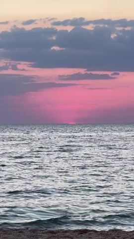The video captures a serene sunset over the sea, likely in Челядиновское сельское поселение, Ukraine. The sky is a vibrant canvas of pink and purple hues, with scattered dark clouds adding depth and texture. The sun, a brilliant pink disc, is partially submerged on the horizon, casting a soft glow across the water. The sea itself is calm, with gentle ripples reflecting the colors of the sky. A small stretch of sandy beach is visible in the foreground, hinting at a coastal setting. The overall mood is peaceful and tranquil, evoking a sense of calm and wonder at the beauty of nature. There is no visible text in the media file.