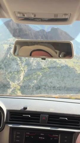 From inside a car driving uphill on a winding road, a glimpse of mountains and a body of water can be seen. The car’s rearview mirror shows the face of a person wearing a light-colored hat. The camera then shifts to capture a broader view of the landscape, revealing a clear, sunny day with mountains leading down to the water, all framed by a rustic wooden fence. The sun shines brightly in the sky.