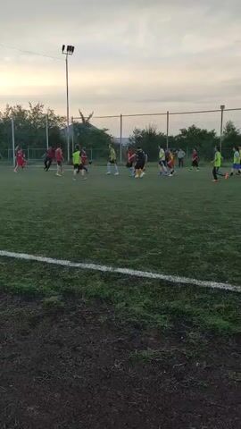 The video shows a youth soccer game on an outdoor artificial turf field. The sky is overcast and the lighting suggests it is late afternoon or early evening. Several children are spread across the field, some in red jerseys and others in yellow or green jerseys, suggesting two teams. Some children are actively engaged with a soccer ball near the center of the field, while others are positioned further away, potentially on defense or offense. A soccer goal is visible on the left side of the frame. A tall light pole with lamps is located behind the players. A net fence runs along the far side of the field. The foreground shows the edge of the field with exposed dirt and grass.