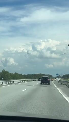 This is a first-person perspective video taken from a vehicle driving on a multi-lane highway. The highway is bordered by a guardrail on the left and trees and vegetation on the right. The sky is partly cloudy with large, white cumulus clouds. In the distance, there are other vehicles on the highway, including a black SUV in front of the viewer's vehicle. There are traffic signs visible on the right side of the highway. The road has clear lane markings, including solid white lines and dashed white lines. The video captures the movement of the vehicle along the highway.