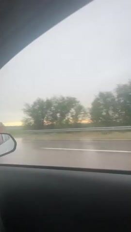 A first-person perspective video shows a car driving on a wet road with a cloudy sky. Raindrops can be seen on the windshield and side window. Another car is visible passing by on the left. The video shows the interior of the car, including the dashboard and a leg of a passenger. The scene appears to be a landscape with trees and a faint sunset in the background.