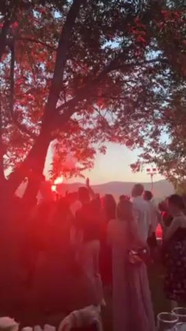 A party crowd of about 20 people is seen from behind, silhouetted against a sunset-tinged sky and lush, rolling hills. The event takes place outdoors, under the canopy of a leafy tree. The scene is lit by a bright red light emanating from handheld flares that are being held by a few individuals, and possibly a spotlight. Many partygoers are holding up their phones to capture the moment. People are casually dressed, and some are holding drinks. A person in white is standing on a raised platform. The atmosphere appears celebratory.