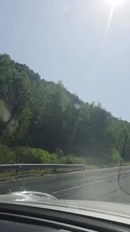 This is a video taken from inside a car, looking forward and slightly to the left. The scene is a road winding through a lush, green, forested hillside under a bright, sunny sky. The sun is high in the sky, suggesting it is midday. The weather appears to be clear and warm.

The road itself is asphalt, with a white line marking the lanes. On the left side of the road, the terrain slopes upwards, covered in dense green foliage, primarily trees and bushes. Some lighter green trees are interspersed with darker, denser foliage, creating a textured appearance. A metal guardrail is visible along the edge of the road on the left for a portion of the video. On the right side of the road, the terrain continues upwards, also covered in trees. The perspective suggests the car is moving along this road, possibly on a scenic route.

There are no people visible in the video. The primary objects are the car's dashboard (partially visible in the foreground), the road, the guardrail, and the natural landscape. The setting is clearly a rural or mountainous area, likely a forest road. The time of day is indicated by the bright sunlight and long shadows. The location context provided is Lozengradtsi, Bulgaria, which fits the description of a forested hillside road. There is no visible text in the video. The overall mood evoked is one of a peaceful drive through nature on a pleasant day.