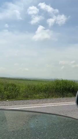 The video shows a view from inside a vehicle driving on a paved road. The road has a white line marking its edge. To the right of the road, there is a grassy field with some scattered bushes and trees. In the distance, there is a hazy line of hills or mountains under a partly cloudy sky. A triangular road sign is visible on a pole to the right of the road in the early frames. A large truck passes by in the opposite direction in the middle of the video. The vehicle appears to be moving at a moderate speed.