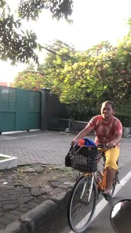 A woman rides an orange bicycle down a paved street. She wears a red t-shirt and yellow pants. A dark-colored bag is in the basket on the front of the bicycle. Trees with green and yellow foliage line the street. A green gate is visible behind the woman. The sun is setting, casting a warm glow on the scene.