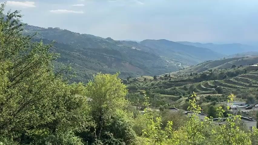 A panoramic video captures a scenic view of a mountainous landscape with lush green vegetation. In the foreground, a dense cluster of trees lines the bottom of the frame. Beyond the trees, rolling hills stretch into the distance, dotted with terraced fields and scattered houses. A road winds through the valley, with several vehicles visible, including cars and trucks. In the upper right portion of the frame, a large power line pylon stands tall against the sky. The sky is partly cloudy, with patches of blue visible. The overall impression is one of a rural, natural setting with signs of human habitation and infrastructure.
