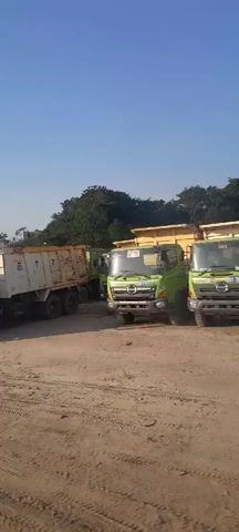 The video shows a line of green dump trucks parked on a dirt lot. Some of the trucks are empty, while others appear to be loaded with dirt. The trucks are parked in a staggered formation, with some facing forward and others angled slightly. The environment is an outdoor lot with a clear blue sky and some trees in the background. The overall impression is that of a construction or industrial site where heavy machinery is stored.
