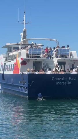 The video shows the stern of a ferry named "Chevalier Paul Marseille" as it moves through calm blue water under a clear sky. The ferry is blue and white with orange accents. Many people are visible on the open-air decks of the ferry, seated and standing, looking towards the camera or out at the water. Some people are leaning on railings, and a few are standing near the entrance to the interior of the ferry. The ferry is creating a visible wake behind it. The water appears clear and reflects the blue sky and the ferry. Other boats and masts of sailboats are visible in the background to the left. The scene suggests a pleasant day for a ferry ride.