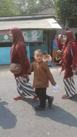 A group of people, including children dressed in traditional attire, are walking down a street. The children are wearing brown striped tops with white and black patterned skirts or pants. Some of the children are holding small Indonesian flags. Adults in the group are wearing maroon headscarves and matching tops, some with white and black patterned skirts. The environment appears to be a street with buildings and trees in the background. The sun is shining, casting shadows on the ground. Some people are watching the procession from the side of the street.