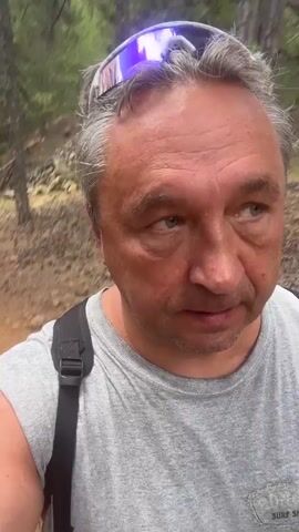 A man with short, graying hair and sunglasses perched on his head is walking outdoors in a wooded area. He is wearing a gray t-shirt and a black backpack. The path appears to be dirt and rocky. The man is looking slightly off-camera to his right.