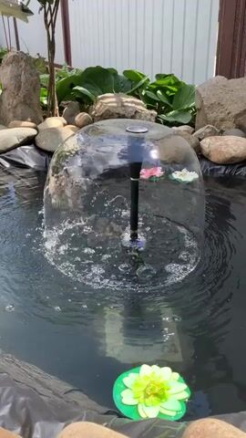 The video shows a close-up of a pond with a solar-powered fountain. The fountain is creating a dome of water. The pond is lined with black plastic and surrounded by rocks and plants. There are also a few decorative items, including a plastic water lily and a yellow rubber duck. In the background, there is a white fence and two decorative bird statues. The lighting suggests it is daytime and sunny.