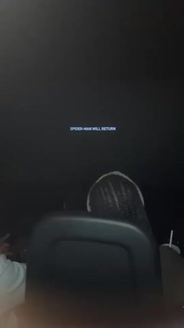 The video begins with a black screen showing white text that reads "SPIDER-MAN WILL RETURN." The image then cuts to a close-up of a light blue object partially covered by white fabric. The focus shifts to a dark interior, possibly a movie theater, with a screen displaying the same "SPIDER-MAN WILL RETURN" text. In the foreground, the backs of two people's heads are visible. The person in the foreground has braided hair, and the person behind them has lighter hair. The environment appears to be dimly lit, typical of a cinema. The video then transitions to another extreme close-up, this time showing a blurred white and blue object, possibly a texture or fabric.