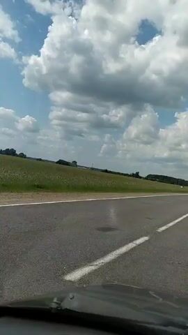 This is a video recording from the perspective of someone driving on a road in a rural area. The scene is dominated by a vast, green field on the left side of the road, stretching towards a distant tree line. The sky above is a bright blue, dotted with large, fluffy white clouds, suggesting a pleasant, possibly sunny day.

The road itself is asphalt and appears to be a two-lane highway with white dividing lines. The perspective shows the road ahead, implying forward motion. The edge of a car's dashboard is visible at the bottom of the frame, confirming the viewpoint is from inside a vehicle.

There are no people or specific objects clearly visible apart from the road, the field, and the sky. The setting appears to be agricultural, with the expansive field being the most prominent feature of the landscape. The time of day seems to be daytime, given the bright sunlight and blue sky. The weather appears to be fair.

No text is visible in the video. The overall mood conveyed is one of travel through a peaceful, open countryside.