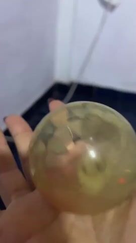 Here's a description of the video:

A hand holds a translucent, yellow, round ball filled with small colored objects. The ball has a textured, almost faceted surface. The hand moves slightly, rotating the ball to show the contents within. A white wall and a dark surface are visible in the background. The lighting is bright, with reflections visible on the surface of the ball. The nails on the hand are painted dark.