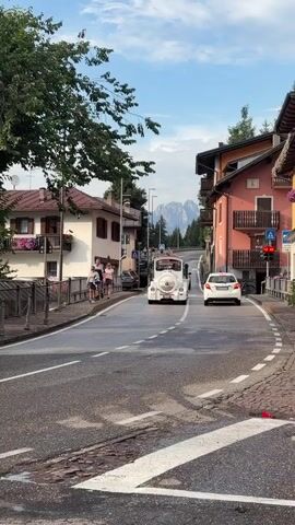 A white tour train is driving down a paved road lined with buildings and trees. Several people are walking on a sidewalk to the left of the road. A white car is driving behind the tour train. A motorcycle is parked on the right side of the road. The sky is blue with some clouds.