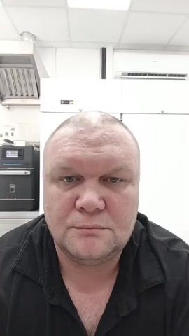 A medium shot video captures a man with a shaved head and a black collared shirt looking directly at the camera. The man's face is the primary focus, with a slightly furrowed brow and a neutral expression. In the background, professional kitchen equipment is visible. To the left, a stainless steel range hood hangs above a dark appliance with a digital display. To the right, a white air conditioning unit is mounted high on the wall. The overall environment appears to be a commercial kitchen setting. The man blinks and shifts his gaze slightly to the left at the 00:04 mark.