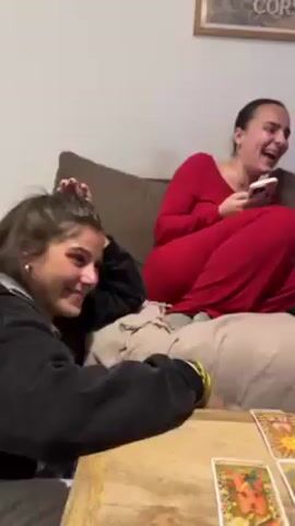 The video shows a group of people in what appears to be a living room. At the beginning, a woman in a red jumpsuit is laughing while looking at her phone. The camera then pans to another woman sitting at a table, covering her mouth with her hand, and looking surprised or emotional. The table has several glasses on it, some filled with glitter. The environment includes a couch, a painting on the wall, and a clothes drying rack. There is a tarot card on a table. The overall mood is lighthearted with moments of surprise and emotion.