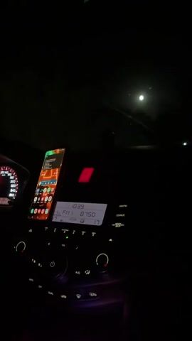 The video shows the interior of a car at night. The dashboard is visible with a smartphone mounted and displaying apps. The car radio display shows FM 1 and a frequency of 87.50. Outside the car, the moon is visible in a dark, cloudy sky. The car appears to be in motion.