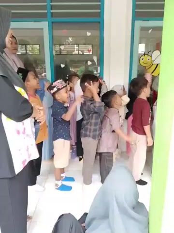 The video shows a group of young children, likely in a kindergarten or preschool setting, standing in a line and looking up at something. Some of the children are pointing. There appear to be teachers or adult supervisors with them. The room has light-colored tile floors and windows, and a cartoon image of a bee is visible on the window. The children are dressed in casual clothing.