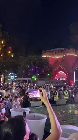At night, a large crowd of people is gathered in front of a brightly lit, castle-like building. Many people in the crowd are holding up their phones, likely to record or take pictures of the event. Some individuals in the crowd are wearing yellow safety vests. In the foreground, there are several white buckets. A person is visible in the foreground, holding up a phone to record. In the center of the frame, a person wearing a yellow safety vest and jeans is standing and appears to be interacting with the crowd, possibly addressing them. Further into the scene, another person is standing on a small platform or stool, also facing the crowd. The building behind them is decorated with numerous colorful lights, creating a festive atmosphere. The sky is dark, indicating it is nighttime. There are trees visible in the background, also decorated with colored lights. The ground in front of the building appears wet, possibly from rain or a water feature.