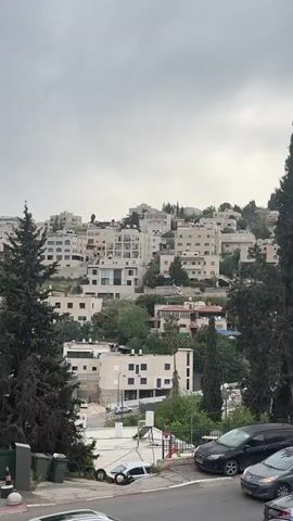 The scene shows a hillside with buildings in a residential area, likely in an urban or suburban setting. The buildings are light-colored, possibly white or beige, and appear to be multi-story dwellings or apartments. The landscape is interspersed with trees, primarily evergreens, which are tall and dense.

In the foreground, there's a street with parked cars on the side, and a green fence or barrier separating the viewer's perspective from a lower area. A glimpse of a white car is also visible.

The sky is overcast, indicating a cloudy or possibly rainy day. The overall impression is that of a moderately dense residential area situated on a slope, with a mix of buildings and natural elements.