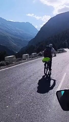 A man on a bicycle rides on a paved road. He is wearing a white shirt, shorts, a hat, and sunglasses. He has a large backpack on the back of his bicycle, with a bright green bag attached to it. The road is bordered by a steep mountain slope on the right, covered with vegetation and rocks. On the left, there is a valley with more mountains in the distance. The sky is clear and blue, with the sun casting a long shadow of the cyclist on the road. The video is shot from a moving perspective, either from another vehicle or a drone, following the cyclist.