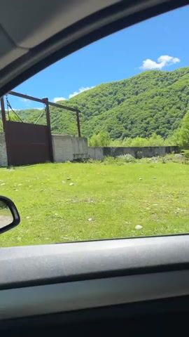 The video offers a view from inside a moving vehicle, looking out through the side window at a sunlit, rural landscape. A concrete fence runs alongside the car's path, bordering a grassy area dotted with scattered rocks. Behind the fence, part of a brick building and a large, rusty metal gate are visible, suggesting an entrance to a property. In the background, lush green mountains, covered in dense foliage, rise under a clear blue sky. The bright sunlight indicates it is daytime. The car appears to be driving past this area. No people or visible text are present in the video. The scene depicts a natural, mountainous environment, consistent with the provided location context of Каменномостский, Russia.