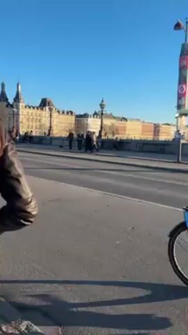 The video shows a bridge on a clear sunny day with people walking and cycling across it. In the background are buildings with ornate architecture. Bicycles pass in the foreground of the video, one has a basket attached to the front.
