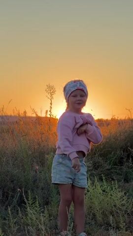 The video features a young girl standing in a field of tall grass and wildflowers at sunset. The sun is low on the horizon, casting a warm, orange glow over the scene. The girl is wearing a pink sweatshirt with a large letter "M" on it, light blue shorts, and a patterned headband. She has a braid in her hair. She appears to be dancing or playing, as she moves her arms and shifts her weight. The surrounding vegetation is lush and green, with some dry stalks of grass visible. In the distance, rolling hills are silhouetted against the vibrant sunset sky. The overall mood is peaceful and idyllic. The time of day is clearly sunset due to the lighting and the color of the sky. The location appears to be a rural or natural setting, given the field of grass and wildflowers. The girl's actions suggest a playful and joyful emotion. There is no visible text in the video.