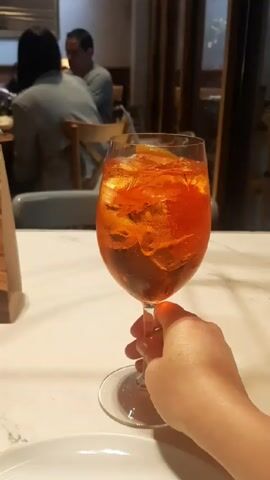 A person's hand is holding a glass of Aperol Spritz on a marble table in a restaurant. The drink has ice and a slice of orange. Other people are sitting at tables in the background.
