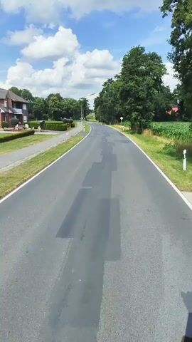 The video shows a first-person perspective of driving down a paved road in a rural area on a sunny day. The road has a white line marking the edge and appears to have some recent patching work on it, indicated by darker rectangular patches. On the left side of the road, there is a sidewalk, a manicured lawn with hedges, and a brick house. Further along the left side, there are trees and streetlights. On the right side of the road, there are tall trees and a field of crops, possibly corn. A speed limit sign indicating 50 is visible on the right side of the road. The sky is blue with scattered white clouds. The overall impression is of a peaceful, scenic drive through the countryside.