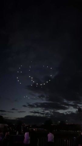The video captures a nighttime scene where a formation of drones creates a swirling, spiral-like shape in the dark sky. The drones are illuminated, appearing as bright dots of light against the night. Below, a crowd of people can be seen observing the display, their silhouettes visible against the dim light. The foreground also includes a metal barrier. Dark clouds are interspersed in the sky, adding texture to the scene. The general environment suggests an outdoor event or gathering.