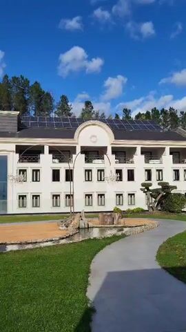 The video pans across a white, multi-story building with solar panels on the roof, set in a landscaped lawn with trees. There's a small pond-like feature, and outdoor chandeliers.  The camera then pans to a view overlooking a small town nestled amongst hills, under a cloudy sky.
