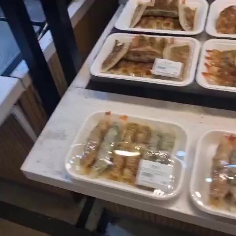 The video shows a display of food items in what appears to be a food stall or a market. In the first frame, a stainless steel surface and a white rectangular object are visible. The subsequent frames reveal food items presented in various ways. There are three white square plates each holding a coiled flatbread-like item with filling. Beneath these, there are white rectangular foam containers. Further down, there are stacks of plastic-wrapped food items that appear to be donuts or pastries. The last frames show bags of what look like dried snacks or chips, one bag containing purple items and several bags containing yellow/orange crispy items. The items are arranged on a white speckled countertop with stainless steel shelving in the background. The lighting is somewhat bright, creating reflections on the metallic surfaces. There are no people directly visible in the video, but the arrangement suggests a commercial setting where food is being sold.