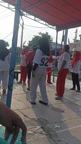 A group of people are gathered outdoors under a red awning. Most of the people are wearing red and white. One person in the center is wearing a baseball jersey with the number 92 on the back. Some people appear to be participating in a game or activity. The ground is paved with concrete. There are trees and buildings in the background. The lighting suggests it is daytime, possibly late afternoon due to the shadows.